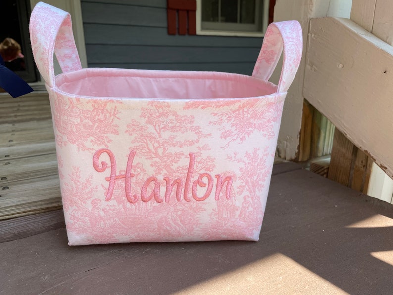 May include: A pink and white fabric basket with handles. The basket features a pink toile pattern and the name "Hanlon" embroidered in pink script. The interior is lined with solid pink fabric.