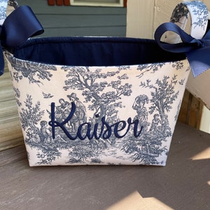 May include: A fabric basket with a blue and white toile pattern featuring a pastoral scene. The basket has navy blue bows on the handles and the name "Kaiser" embroidered in navy blue.