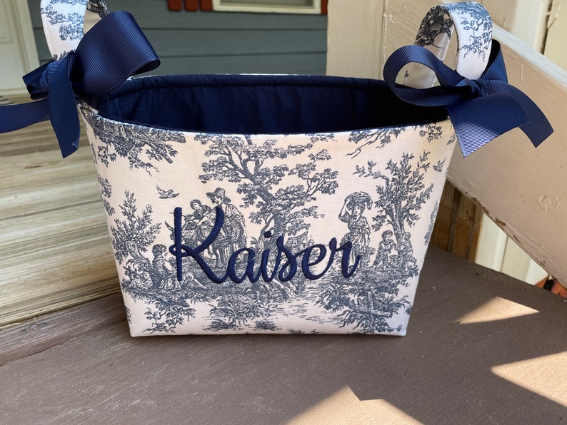 May include: A fabric basket with a blue and white toile pattern featuring a pastoral scene. The basket has navy blue bows on the handles and the name "Kaiser" embroidered in navy blue.