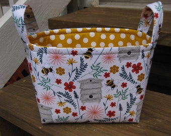 May include: A white fabric storage basket with yellow polka dot lining. The basket has two handles and features a bee and flower pattern.