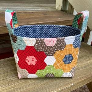 May include: A handmade fabric basket with a patchwork design. The exterior features a variety of hexagonal fabric pieces in floral and geometric patterns, including red, green, and brown. The interior is lined with a blue patterned fabric, and it has two fabric handles.