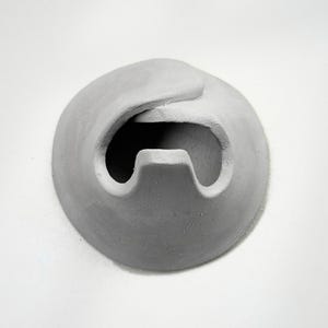 May include: A gray, abstract sculpture with a rounded shape and a curved opening. The sculpture has a textured surface and appears to be made of a solid material. The design is simple and modern.