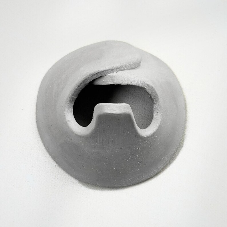 May include: A gray, abstract sculpture with a rounded shape and a curved opening. The sculpture has a textured surface and appears to be made of a solid material. The design is simple and modern.