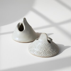 May include: Two ceramic vessels with a handle and a circular opening. One is smooth, the other has a textured, topographic pattern. Both are off-white and sit on a white surface, with shadows.