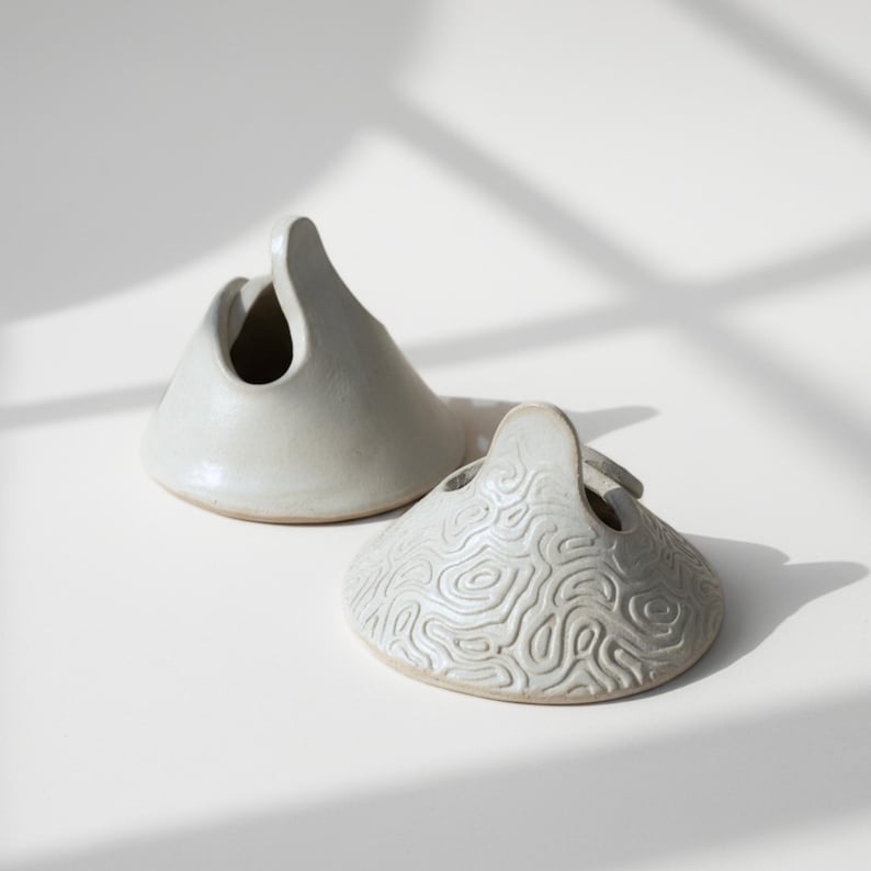 May include: Two ceramic vessels with a handle and a circular opening. One is smooth, the other has a textured, topographic pattern. Both are off-white and sit on a white surface, with shadows.