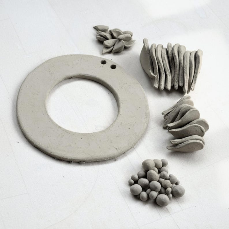 Work-in-progress shot of clay wreath base, leaves, and berries laid out with holes for hanging, demonstrating slab-building pottery kit components and pottery tools.