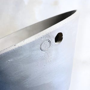 May include: Close-up of a light grey, concrete planter with a rounded oval indentation and a circular hole. The planter has a smooth, curved design and a textured surface. The background is a soft, neutral colour.