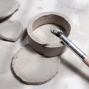 May include: A close-up shot of pottery in progress. A light gray clay ring and a circular base are visible, with a paintbrush resting on the ring. A heart-shaped clay piece is also present. The clay has visible texture from the artist's tools.
