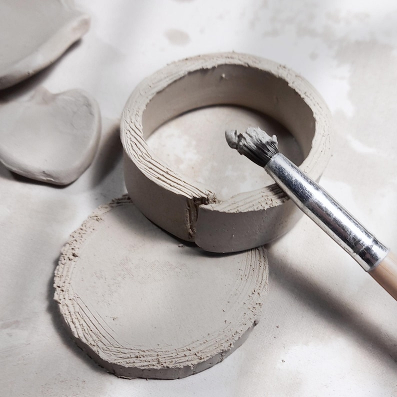 May include: A close-up shot of pottery in progress. A light gray clay ring and a circular base are visible, with a paintbrush resting on the ring. A heart-shaped clay piece is also present. The clay has visible texture from the artist's tools.