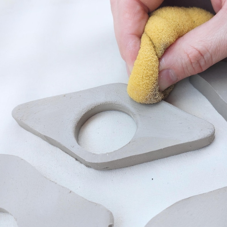 May include: A hand using a yellow sponge to smooth the surface of a gray, diamond-shaped ceramic piece with a circular hole. Other similar pieces are visible in the background, suggesting a crafting or pottery project.