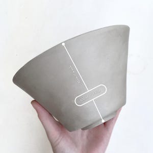 Close-up of hand holding handbuilt bowl, leather-hard clay with visible grog and sponge marks under natural light. Details show authenticity and handmade character.