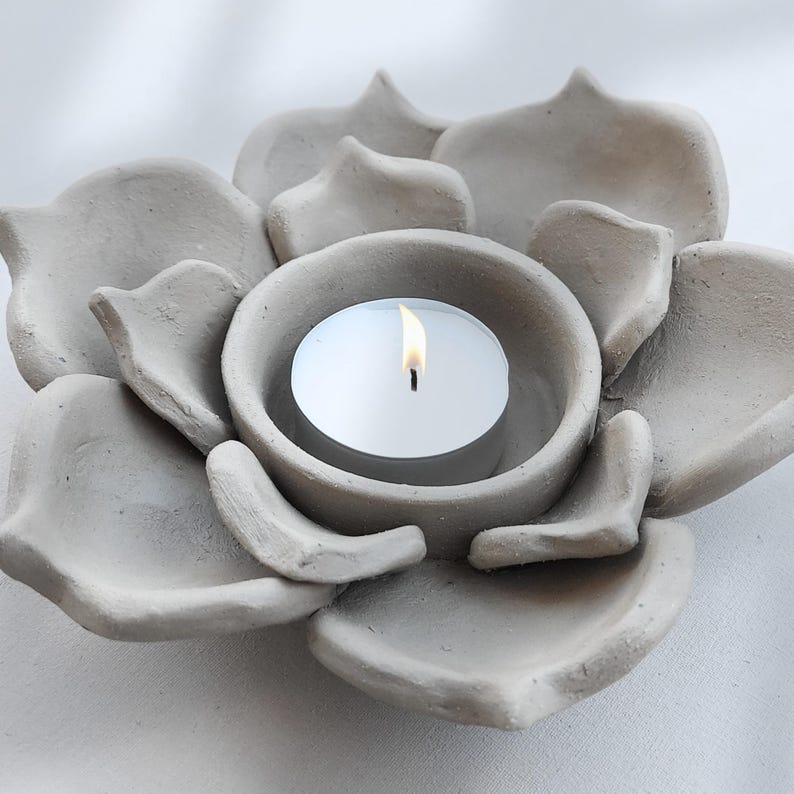 May include: A gray, flower-shaped candle holder with a lit white tea light candle. The candle holder is made of a textured, concrete-like material. The petals of the flower are arranged around a central cup that holds the candle.