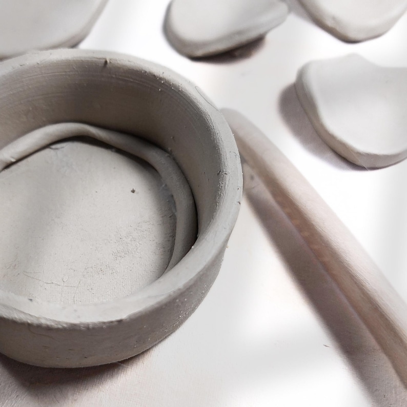 May include: A close-up of pottery tools and clay pieces. A small, round, gray clay bowl is in the foreground, with a wooden tool and several flat, oval clay pieces in the background. The items are arranged on a white surface.