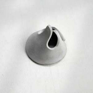 May include: A small, gray, ceramic sculpture with a rounded base and a curved, open top. The sculpture has a smooth, matte finish and appears to be handmade. The design is abstract and organic, resembling a stylized vessel or a folded form.