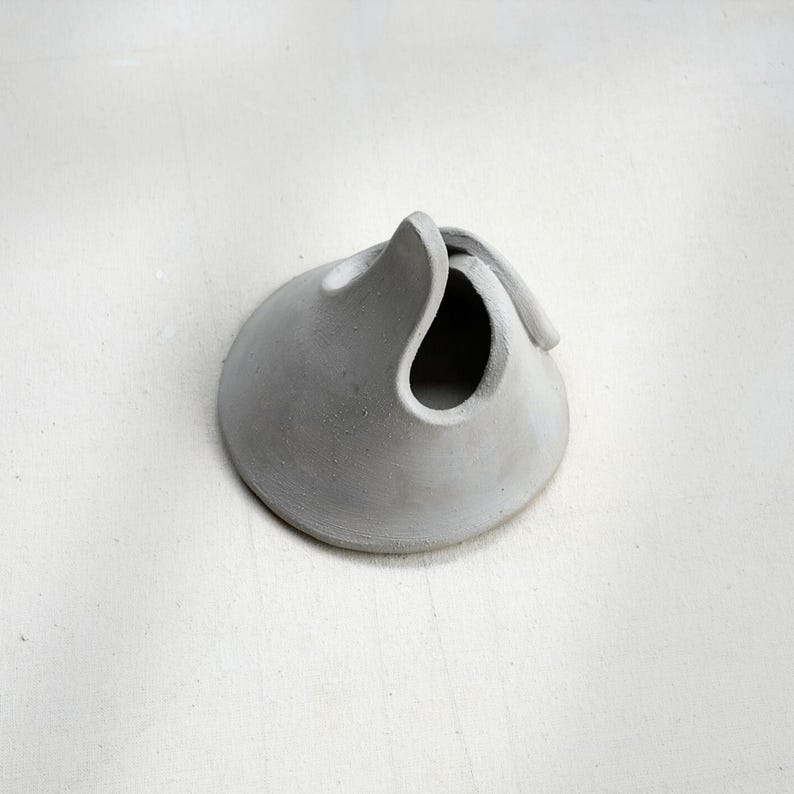 May include: A small, gray, ceramic sculpture with a rounded base and a curved, open top. The sculpture has a smooth, matte finish and appears to be handmade. The design is abstract and organic, resembling a stylized vessel or a folded form.