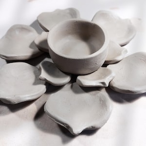 May include: A collection of handmade, unglazed ceramic pieces. The arrangement includes a small, round bowl and several petal-shaped dishes in varying sizes. The neutral color and organic shapes suggest a minimalist aesthetic, suitable for small items or decorative use.