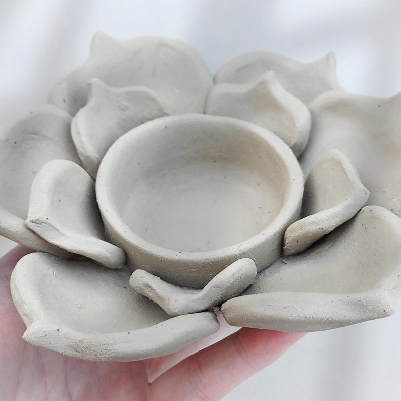 May include: A handmade, light gray ceramic bowl shaped like a flower. The bowl has a central circular depression surrounded by petal-like structures. The piece is held in a hand, showcasing its size and texture.