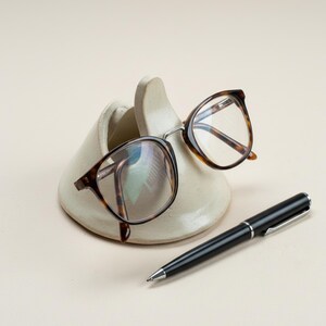 May include: A pair of tortoiseshell eyeglasses rests on a cream-colored ceramic glasses holder. A black pen with silver accents lies beside the holder. The eyeglasses have a classic, round frame.