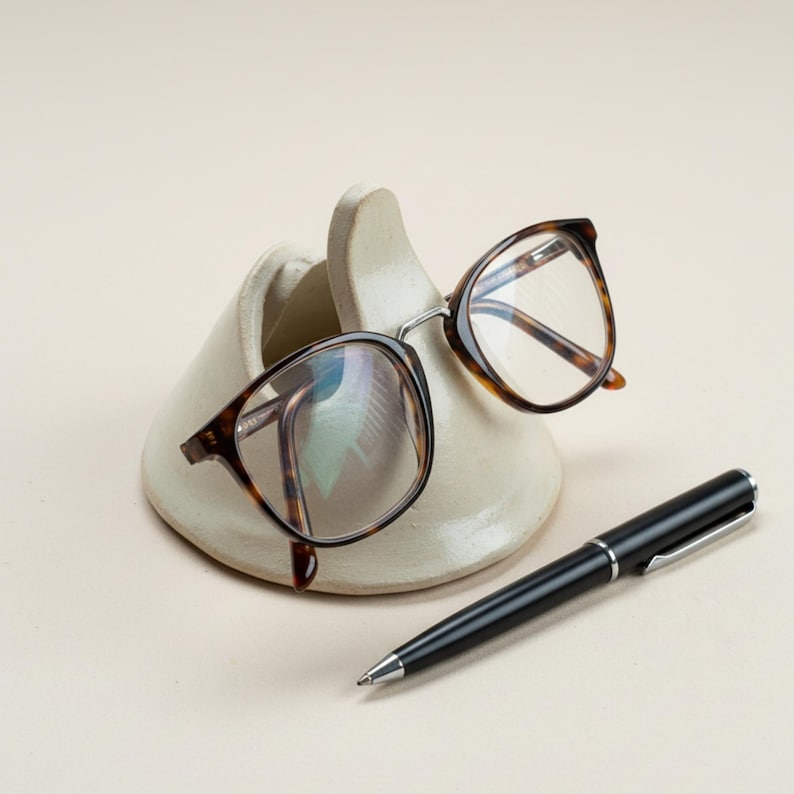 May include: A pair of tortoiseshell eyeglasses rests on a cream-colored ceramic glasses holder. A black pen with silver accents lies beside the holder. The eyeglasses have a classic, round frame.