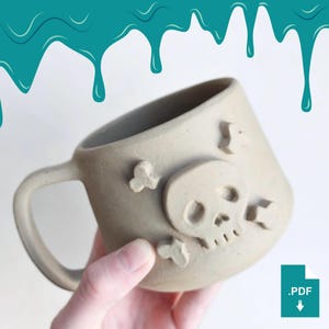 Può includere: Una tazza in ceramica beige con un design a teschio e ossa incrociate. La tazza ha un manico ed è decorata con un teschio in rilievo e due ossa incrociate. La tazza è tenuta da una mano.
