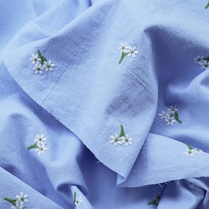 Pode incluir: Tecido azul claro com bordados de flores brancas e caules verdes. O tecido é macio e ligeiramente enrugado, com bordados florais distribuídos uniformemente pela superfície. A estética geral é delicada e fresca.