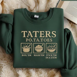 Puede incluir: Sudadera verde bosque con la inscripción dorada "TATERS P.O.TA.TOES". Debajo, tres ilustraciones de platos de patatas. El texto debajo dice "BOIL'EM MASH'EM STICK'EM IN A STEW".