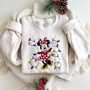 Op de afbeelding: Cr&egrave;mekleurige sweatshirt met een geborduurd Minnie Mouse-ontwerp. Minnie draagt een rode polkadotjurk en houdt een slinger van kleurrijke kerstverlichting vast. De sweatshirt heeft lange mouwen en een ronde hals.