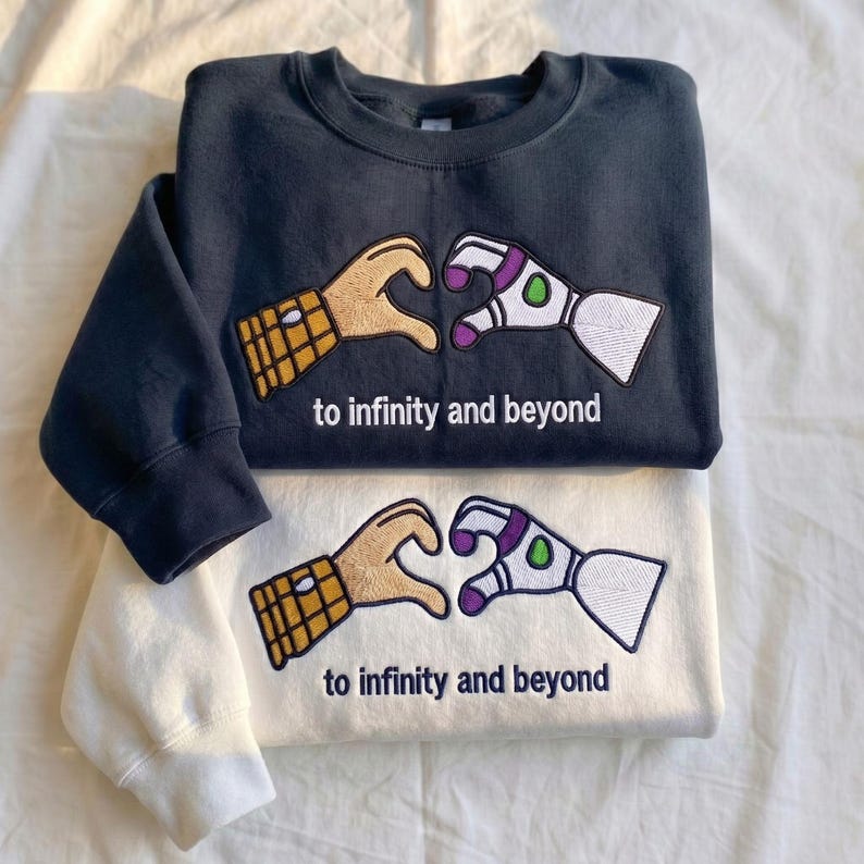 Puede incluir: Se muestran dos sudaderas, una azul marino y otra blanquecina. Cada una presenta un dise&ntilde;o bordado de dos manos formando un coraz&oacute;n, una que se asemeja a un guante y la otra al brazo de Buzz Lightyear, con el texto "to infinity and beyond" debajo.