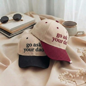 Puede incluir: Dos gorras de béisbol beige con la frase bordada "go ask your dad". Una gorra tiene la visera negra, la otra burdeos. Las gorras están apiladas una encima de la otra. Gafas de sol y una pila de libros en el fondo.