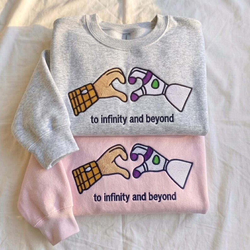 Puede incluir: Dos sudaderas, una gris y otra rosa, est&aacute;n apiladas. Cada una presenta un dise&ntilde;o bordado de dos manos formando un coraz&oacute;n, una un guante marr&oacute;n y la otra una mano de traje espacial blanca y morada. El texto "to infinity and beyond" est&aacute; debajo.