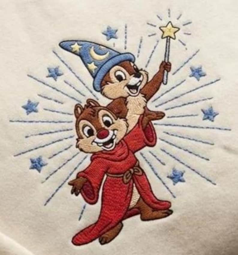 May include: Embroidered design featuring two cartoon chipmunks. One chipmunk wears a red robe and the other a blue wizard hat with stars and a crescent moon. The chipmunk in the hat holds a star-tipped wand. Blue starburst and stars surround the chipmunks.
