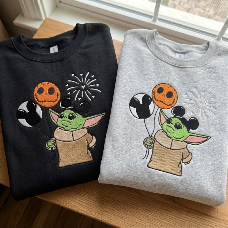 May include: A black sweatshirt and a grey sweatshirt, each featuring an embroidered design of a green creature with large ears wearing mouse-ear headbands, holding a pumpkin-faced balloon and a black and white balloon, with fireworks above.