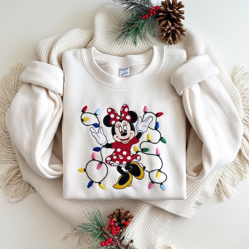 Op de afbeelding: Cr&egrave;mekleurige sweatshirt met een geborduurd Minnie Mouse-ontwerp. Minnie draagt een rode polkadotjurk en houdt een slinger van kleurrijke kerstverlichting vast. De sweatshirt heeft lange mouwen en een ronde hals.
