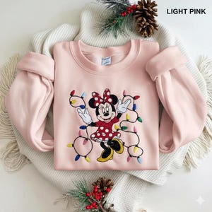 Op de afbeelding: Lichtroze sweatshirt met een geborduurd Minnie Mouse-ontwerp en kerstverlichting. Minnie Mouse draagt een rode jurk met witte stippen en een bijpassende strik. De tekst "LIGHT PINK" staat rechtsboven.