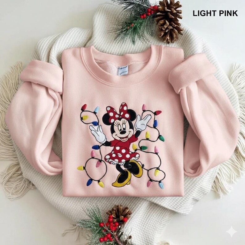 Op de afbeelding: Lichtroze sweatshirt met een geborduurd Minnie Mouse-ontwerp en kerstverlichting. Minnie Mouse draagt een rode jurk met witte stippen en een bijpassende strik. De tekst "LIGHT PINK" staat rechtsboven.