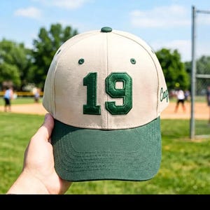 Puede incluir: Gorra de béisbol con corona color crema y visera verde. El número "19" está bordado en verde en la parte delantera. La gorra se sostiene frente a un campo de béisbol.