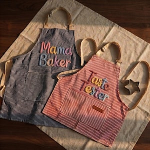 Op de afbeelding: Twee schorten, een marineblauw gestreept met de tekst "Mama Baker" in kleurrijke letters, en een rood gestreept met "Taste Tester". Beide hebben zakken en beige bandjes. Een stervormige koekjesvorm is ook zichtbaar.