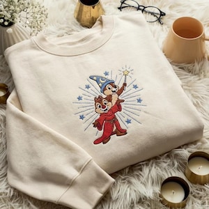 May include: Cream-colored sweatshirt featuring an embroidered design of two cartoon chipmunks, one wearing a wizard hat and holding a star wand. The design is surrounded by blue starbursts and stars.