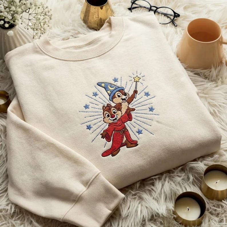 May include: Cream-colored sweatshirt featuring an embroidered design of two cartoon chipmunks, one wearing a wizard hat and holding a star wand. The design is surrounded by blue starbursts and stars.