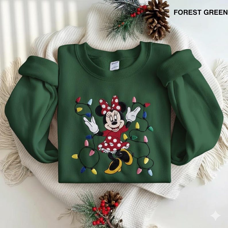 Op de afbeelding: Bostgroen sweatshirt met een geborduurd Minnie Mouse-ontwerp. Minnie draagt een rode jurk met witte stippen en een bijpassende strik. Ze is omringd door kleurrijke kerstverlichting. De tekst "FOREST GREEN" staat rechtsboven.