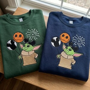 May include: A dark green and a navy blue crewneck sweatshirt, each featuring an embroidered graphic of a green creature with large ears holding three balloons: one black and white Mickey-shaped, one orange pumpkin-faced, and white fireworks.