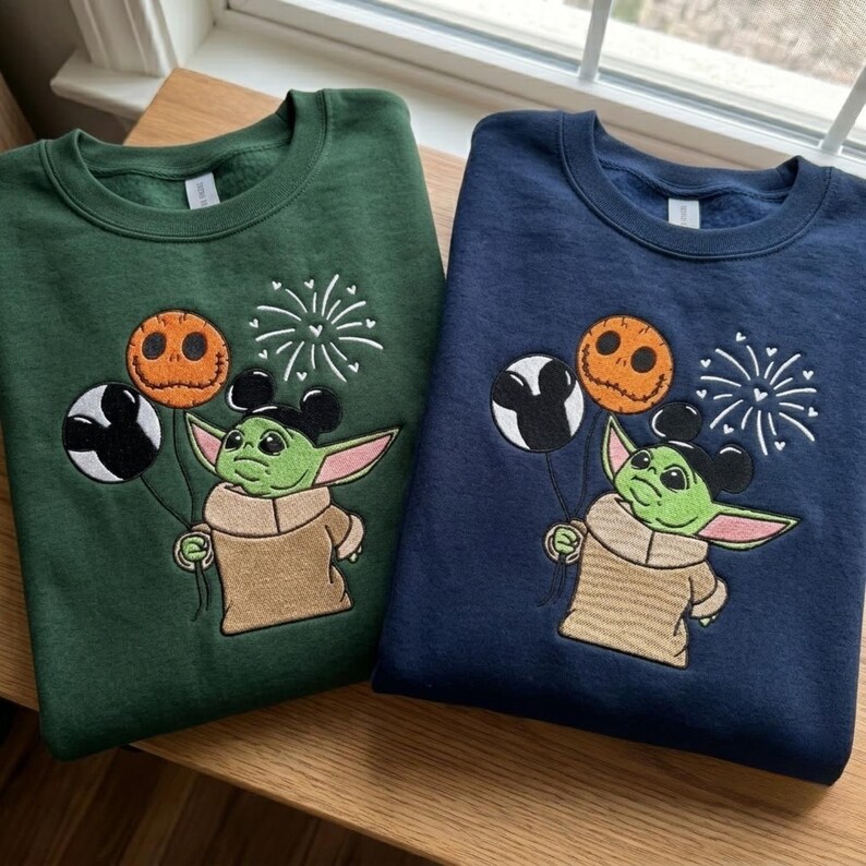 May include: A dark green and a navy blue crewneck sweatshirt, each featuring an embroidered graphic of a green creature with large ears holding three balloons: one black and white Mickey-shaped, one orange pumpkin-faced, and white fireworks.