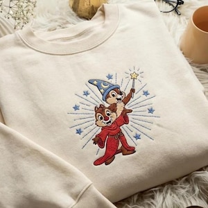 May include: Cream-colored sweatshirt featuring an embroidered design of two cartoon chipmunks. One chipmunk wears a wizard hat and holds a wand with a star, while the other wears a red robe. The design is surrounded by blue starbursts.