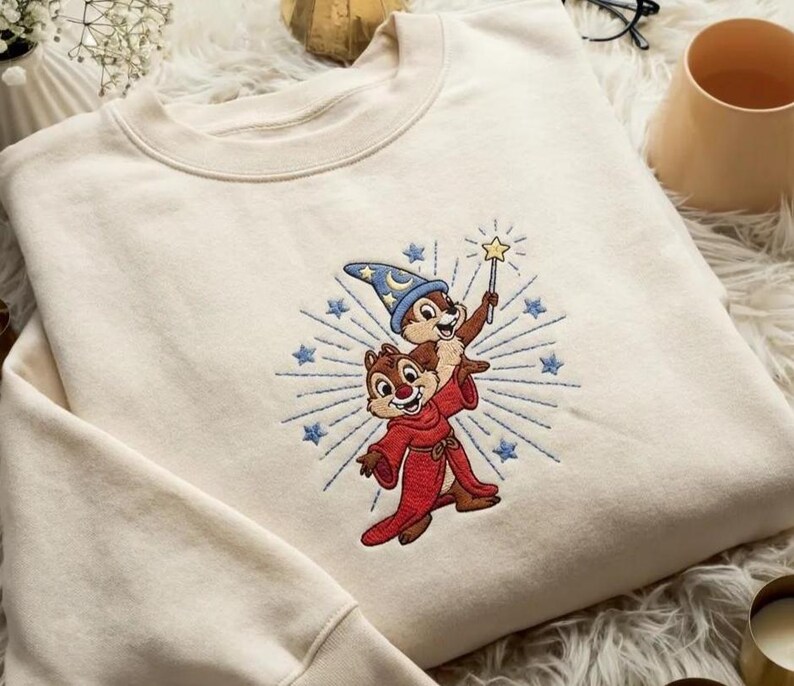 May include: Cream-colored sweatshirt featuring an embroidered design of two cartoon chipmunks. One chipmunk wears a wizard hat and holds a wand with a star, while the other wears a red robe. The design is surrounded by blue starbursts.