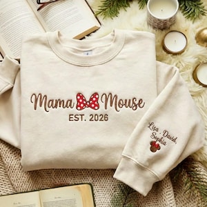 Puede incluir: Sudadera color crema con el texto bordado "Mama Mouse" y "EST. 2026". Un lazo de lunares rojos y blancos está presente en el diseño. La manga tiene los nombres "Lisa, David, Sophia" bordados.