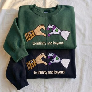 Puede incluir: Dos sudaderas, una verde oscuro y otra negra, apiladas. Cada una presenta un dise&ntilde;o bordado de dos manos formando un coraz&oacute;n, una que se asemeja a un guante y la otra a un brazo rob&oacute;tico. El texto "to infinity and beyond" est&aacute; debajo.