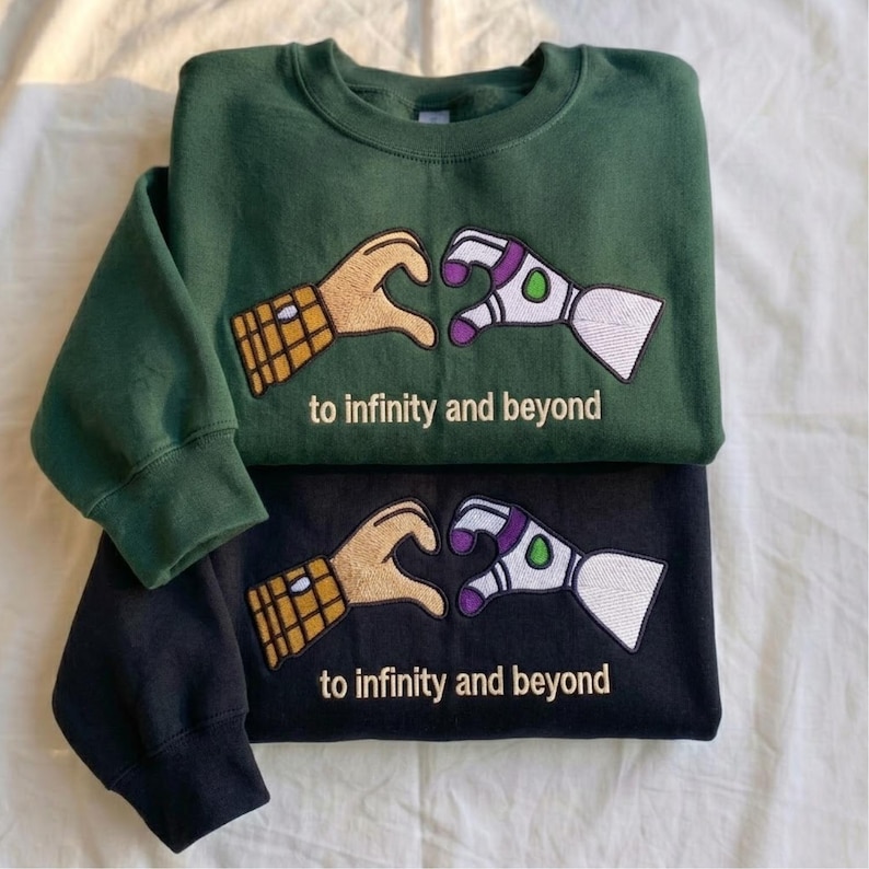 Puede incluir: Dos sudaderas, una verde oscuro y otra negra, apiladas. Cada una presenta un dise&ntilde;o bordado de dos manos formando un coraz&oacute;n, una que se asemeja a un guante y la otra a un brazo rob&oacute;tico. El texto "to infinity and beyond" est&aacute; debajo.