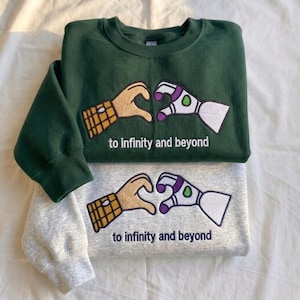 Puede incluir: Dos sudaderas, una verde bosque y otra gris jaspeado, apiladas. Cada una presenta un dise&ntilde;o bordado de dos manos formando un coraz&oacute;n, una con un guante amarillo y marr&oacute;n, la otra un brazo rob&oacute;tico blanco y morado. El texto "to infinity and beyond" est&aacute; debajo.