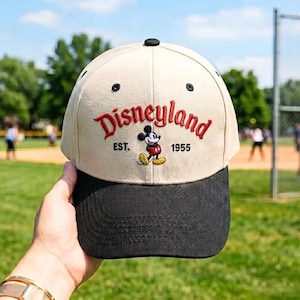 Op de afbeelding: Een beige en zwarte baseballpet met het woord "Disneyland" geborduurd in rood script. De pet heeft een Mickey Mouse-afbeelding en de tekst "EST. 1955". De pet heeft een zwarte klep en een zwarte knop aan de bovenkant.