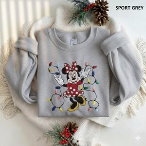 Op de afbeelding: Sportgrijs sweatshirt met een geborduurd Minnie Mouse-ontwerp. Minnie draagt een rode jurk met witte stippen en een bijpassende strik. Ze is omringd door kleurrijke kerstverlichting. De tekst "SPORT GREY" staat rechtsboven.